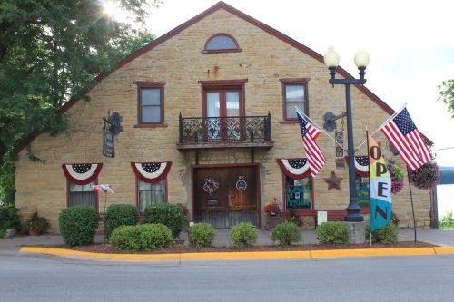 Historic stone building with red trim, flags, and an "OPEN" sign, surrounded by greenery and a view of the water.