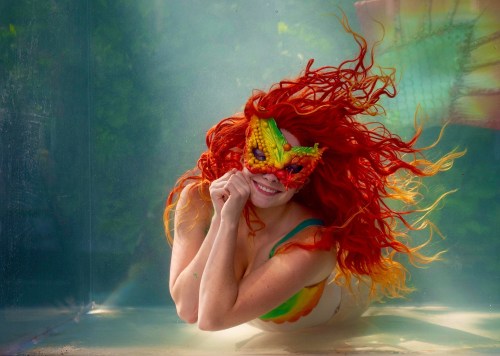 A woman with vibrant red hair and a colorful mask swims underwater, surrounded by a shimmering blue backdrop.