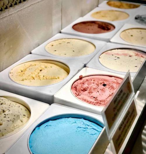 A variety of colorful ice cream flavors in tubs, including blue, pink, yellow, and brown, displayed in a freezer.