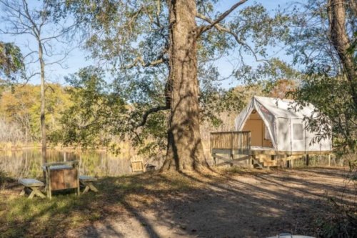 A glamping tent by a serene lake, surrounded by trees and a clear blue sky.