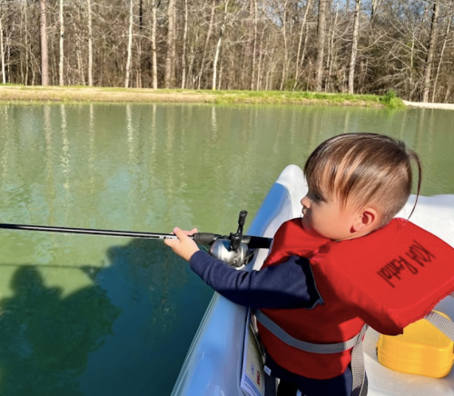 A young child in a life jacket fishing from a small boat on a calm, greenish water body surrounded by trees.