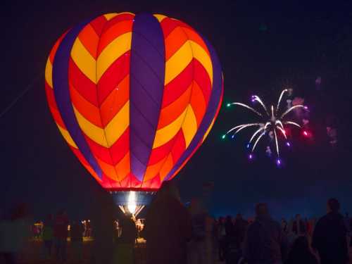 A brightly colored hot air balloon glows at night, with fireworks bursting in the sky above a crowd of spectators.