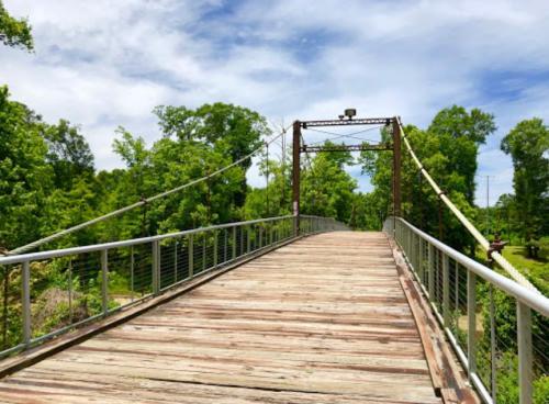 A wooden suspension bridge surrounded by lush green trees under a blue sky with scattered clouds.
