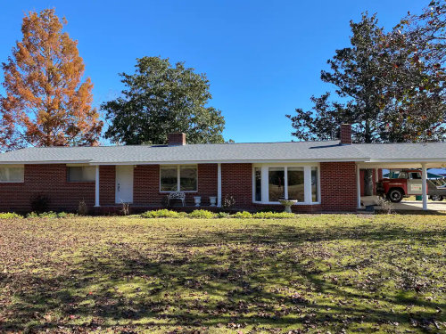 A single-story brick house with a front porch, surrounded by trees and a lawn covered in fallen leaves.