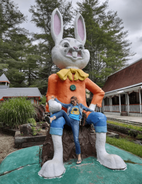 A person poses playfully beside a large, colorful statue of a cartoon rabbit in a park setting.