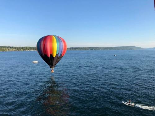 A colorful hot air balloon floats above a calm lake, with boats visible in the distance under a clear blue sky.