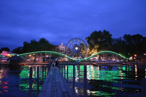 A vibrant amusement park at dusk, featuring a roller coaster and a ferris wheel, reflected in calm water.
