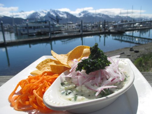 A plate with creamy dip, tortilla chips, and carrot ribbons, set against a scenic marina with mountains in the background.