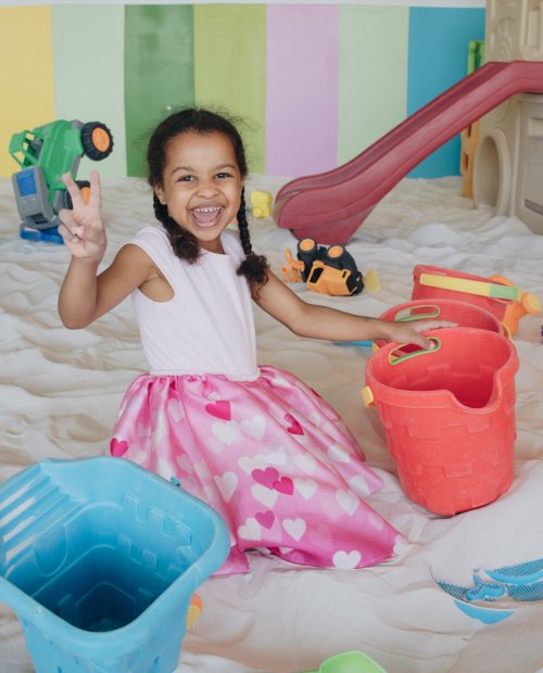 A joyful girl in a pink heart-patterned skirt plays in a colorful sandbox with toy buckets and a slide in the background.