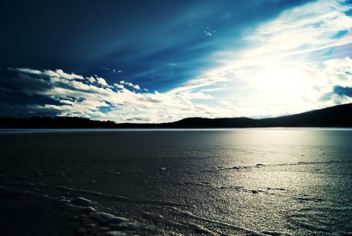 A serene winter landscape with a frozen lake, dramatic clouds, and sunlight reflecting off the icy surface.