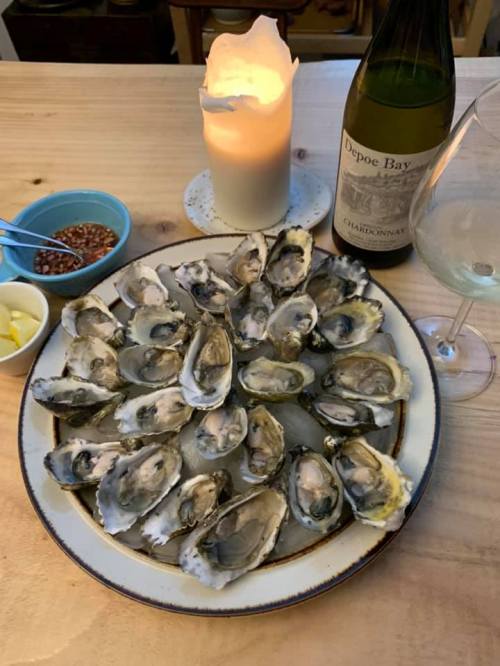 A platter of fresh oysters on a table, accompanied by a candle, wine, and condiments.