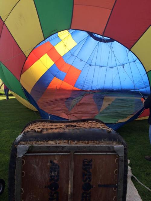 A colorful hot air balloon partially inflated, with a wicker basket in the foreground on green grass.