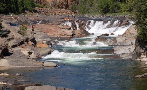 A scenic river with cascading waterfalls surrounded by rocky terrain and lush greenery, with people enjoying the area.