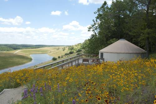A scenic view of a yurt surrounded by wildflowers, overlooking a river and rolling hills under a blue sky.