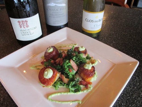 A plate of seared scallops on a bed of kale, garnished with sauce, alongside three wine bottles.