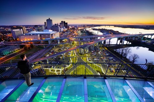 A person stands on a glass observation deck overlooking a city skyline at sunset, with rivers and highways visible below.