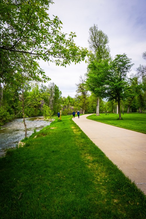 A scenic park path alongside a stream, with people walking among lush green trees and grass under a cloudy sky.