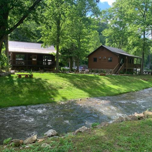 A serene scene of two cabins by a flowing stream, surrounded by lush green trees and grass.