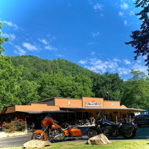 A motorcycle shop with two parked bikes in front, surrounded by greenery and mountains under a blue sky.