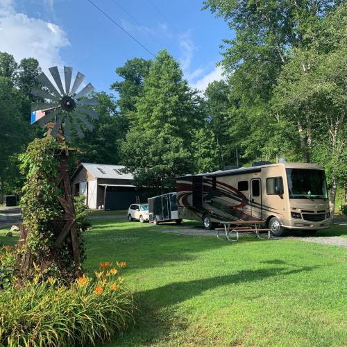 A recreational vehicle parked near a windmill and a barn, surrounded by trees and greenery on a sunny day.