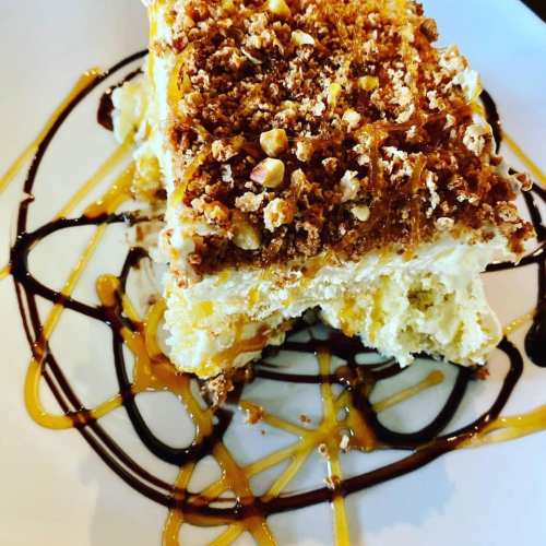 A slice of dessert topped with crushed nuts, drizzled with chocolate and caramel sauces on a white plate.