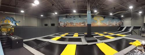 A spacious indoor trampoline park featuring multiple trampolines with black and yellow mats and a colorful mural on the wall.