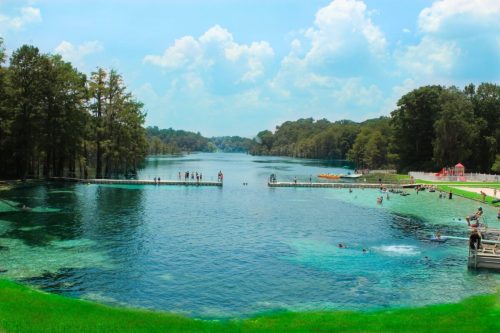 A scenic view of a clear blue river surrounded by trees, with people swimming and enjoying the water on docks.