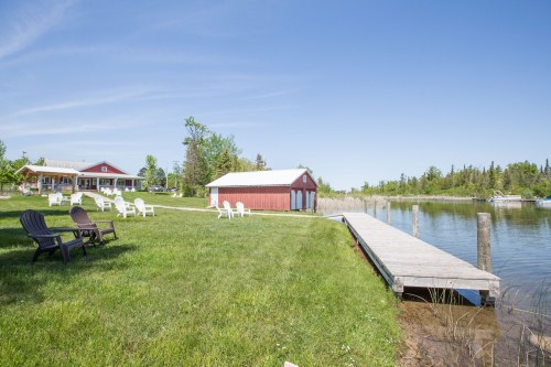 A serene lakeside scene featuring a red shed, dock, and white chairs on green grass under a clear blue sky.