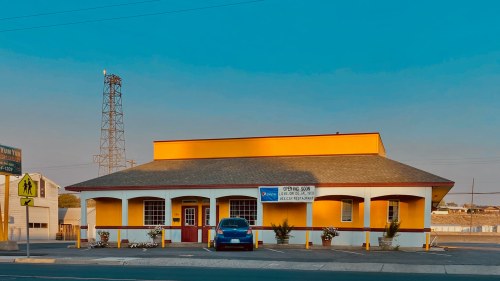 A bright yellow building with a red door and windows, featuring a sign that says "Opening Soon." A blue car is parked outside.
