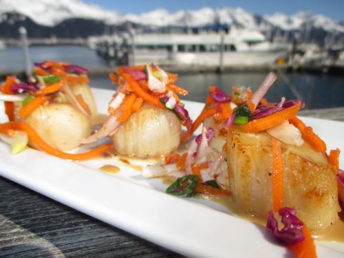 Three seared scallops topped with colorful vegetable slaw, set against a scenic waterfront and snowy mountains.