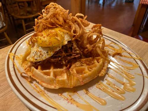 A waffle topped with pulled pork, a fried egg, crispy onions, and drizzled with sauce on a wooden table.