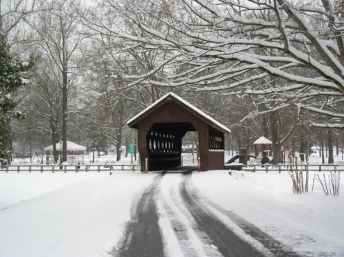 A snow-covered road leads to a wooden covered bridge surrounded by bare trees and a winter landscape.