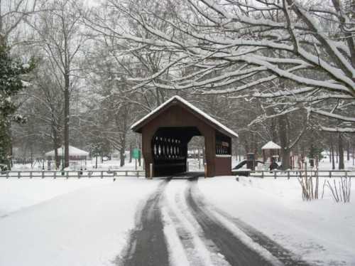 A snow-covered road leads to a wooden covered bridge surrounded by bare trees and a winter landscape.