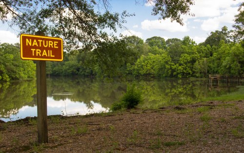 A sign reading "Nature Trail" near a calm lake surrounded by lush greenery and trees under a partly cloudy sky.