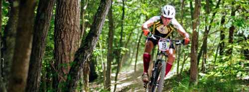 A mountain biker navigates a wooded trail, focused and in motion, wearing a helmet and racing gear.