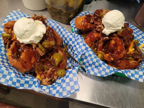 Two loaded BBQ sandwiches topped with jalapeños, barbecue sauce, and a dollop of whipped cream on blue checkered paper.
