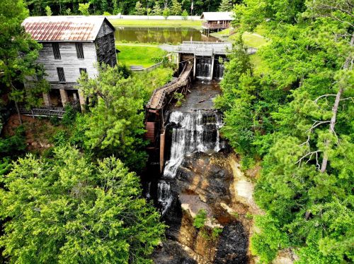 Aerial view of a rustic mill beside a waterfall, surrounded by lush greenery and a calm pond.