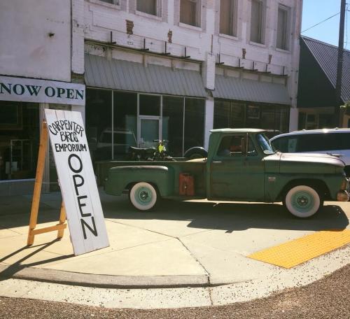 A vintage green pickup truck parked outside a shop with a sign reading "Carpenter Bros Emporium - OPEN."