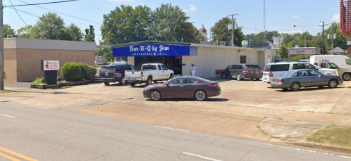 A barbecue restaurant with parked cars in front, surrounded by trees and power lines on a sunny day.