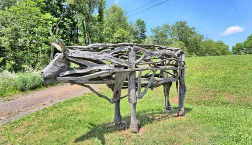 A wooden sculpture of a cow made from intertwined branches, set in a grassy area with trees in the background.