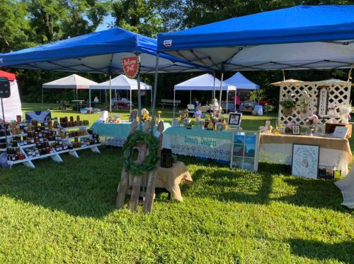 A vibrant outdoor market scene with blue tents displaying various handmade goods and jars on tables, surrounded by greenery.
