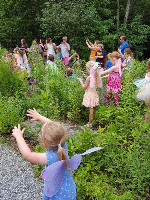 A group of children in colorful outfits play and dance in a lush green garden, some wearing fairy wings.