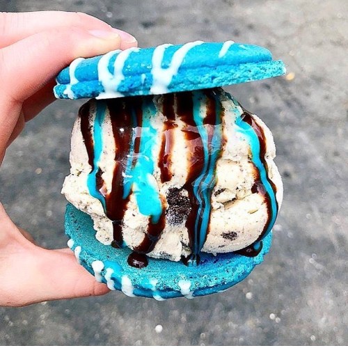 A hand holding a vibrant blue cookie sandwich filled with ice cream and drizzled with chocolate sauce.