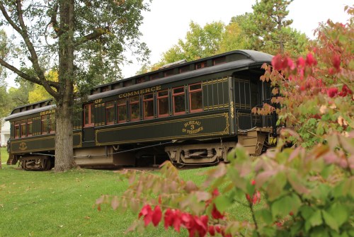 A vintage train car, painted in dark green and gold, sits among colorful autumn foliage in a park setting.