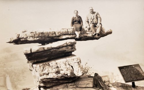 A man and woman sit on a large rock formation, with a scenic view and informational sign nearby.