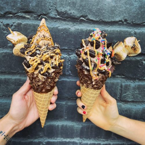 Two hands hold large ice cream cones topped with chocolate, sprinkles, and toasted marshmallows against a dark background.
