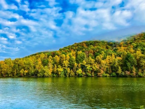 A serene lake surrounded by vibrant autumn trees and a cloudy blue sky reflecting on the water's surface.