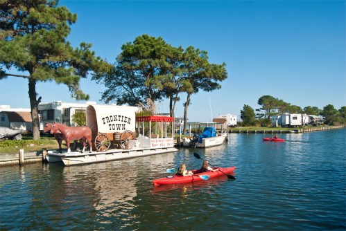 Two kayakers paddle on a calm waterway beside a vintage wagon and trees, with RVs in the background under a clear blue sky.