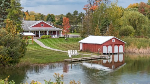 A red house and boathouse by a calm lake, surrounded by lush greenery and autumn-colored trees.
