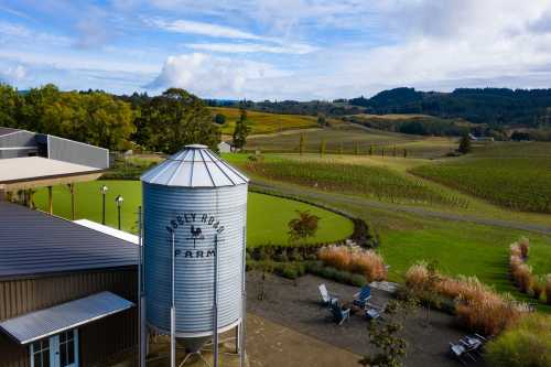 A scenic view of a farm with a silos, vineyards, and rolling hills under a partly cloudy sky.